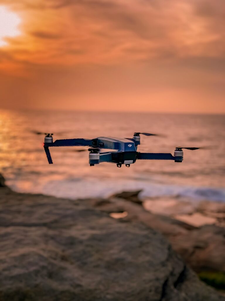 Home A drone hovers over a rocky shoreline with a stunning sunset backdrop, creating a serene atmosphere.